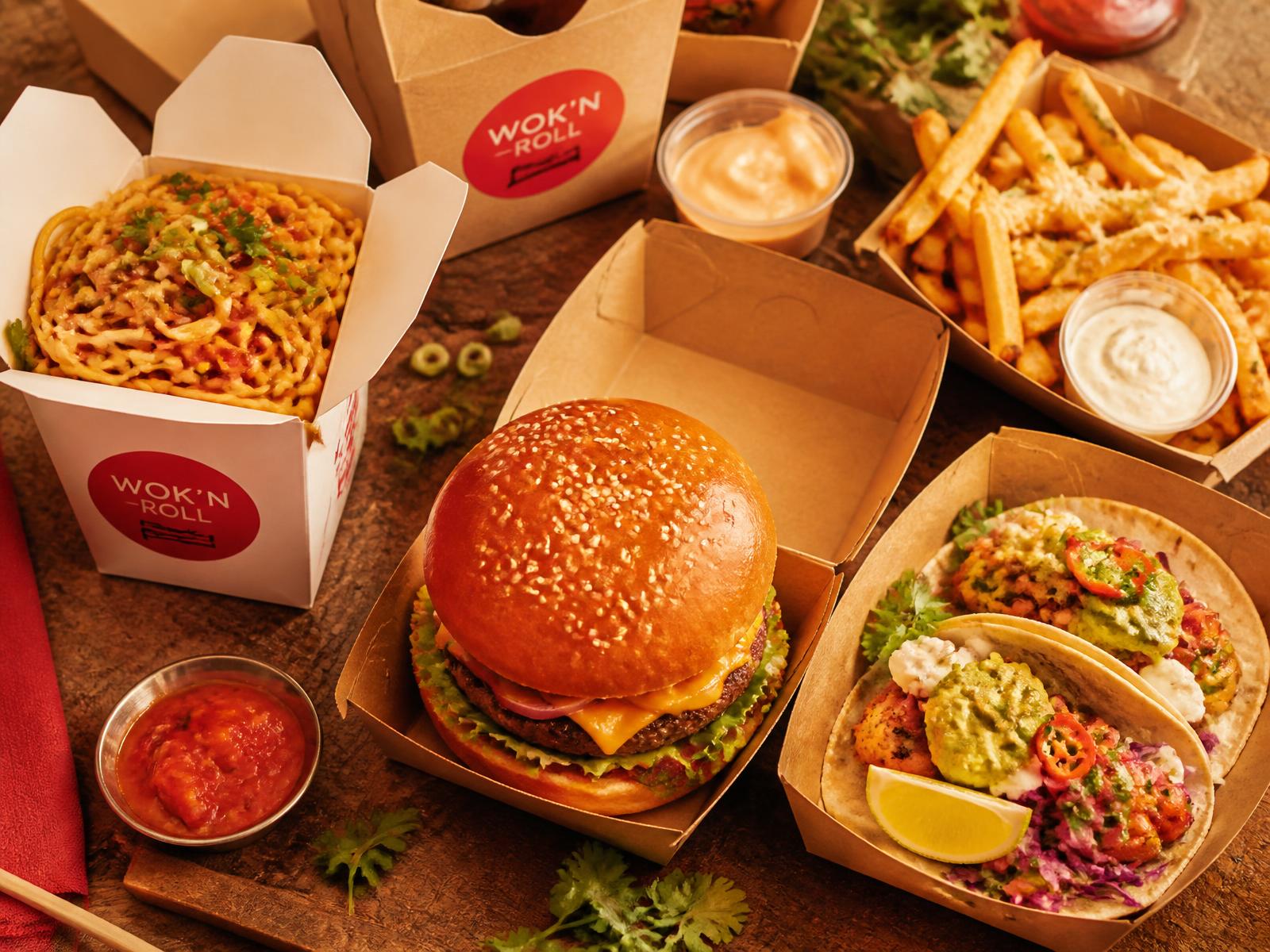 Takeout containers with noodles, a burger, tacos, and fries on a wooden table